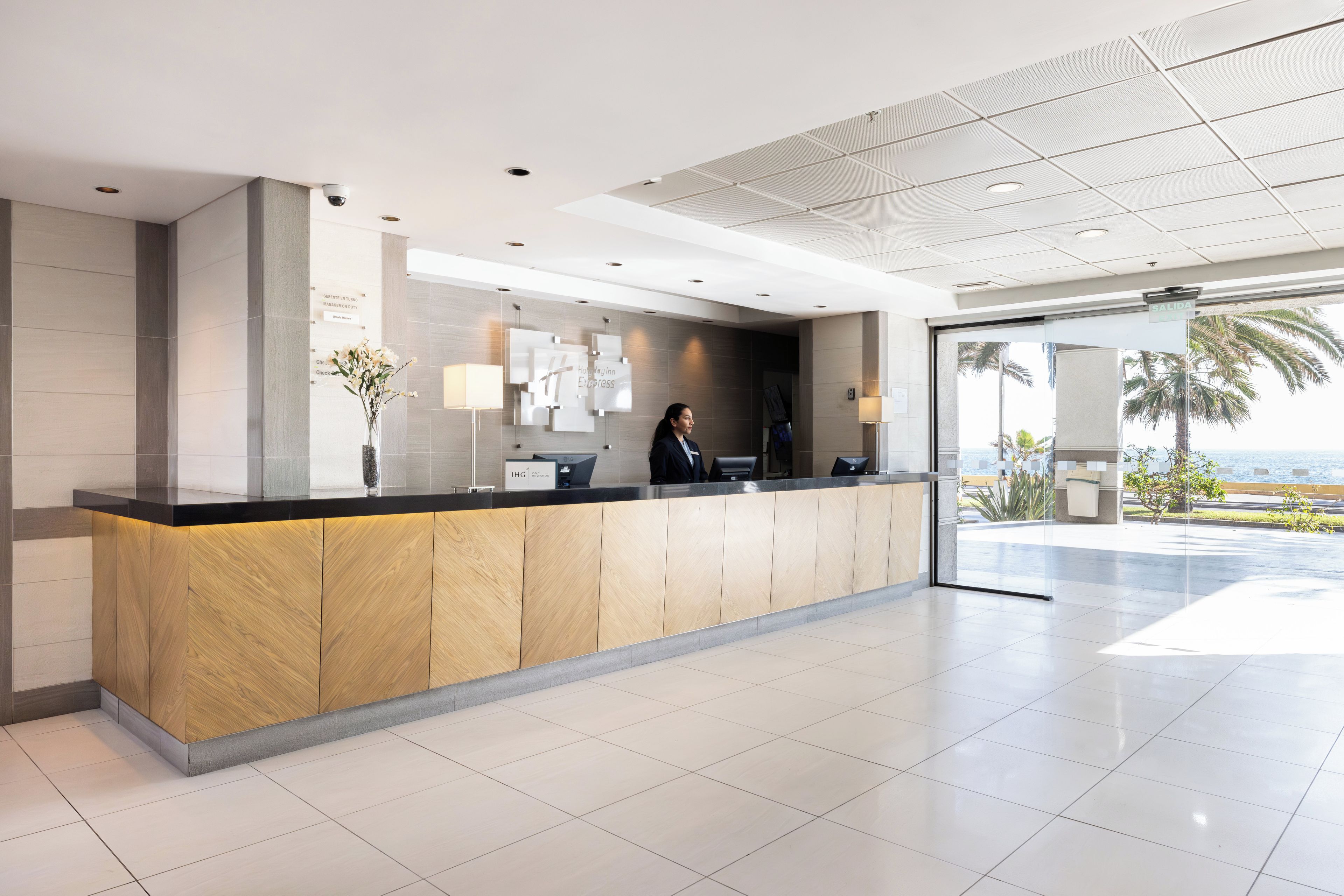 A modern reception desk with a receptionist, a digital check-in station, and a view of the ocean through a glass door.