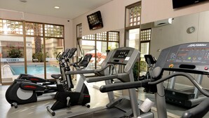 Une salle de sport équipée de tapis de course, de vélos elliptiques et dotée d’un espace piscine visible à travers de grandes fenêtres.