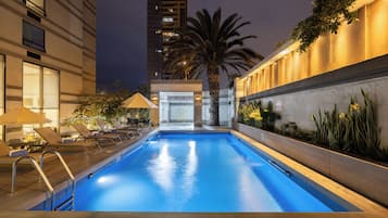 A swimming pool with sun loungers and a palm tree, surrounded by a building with illuminated windows.