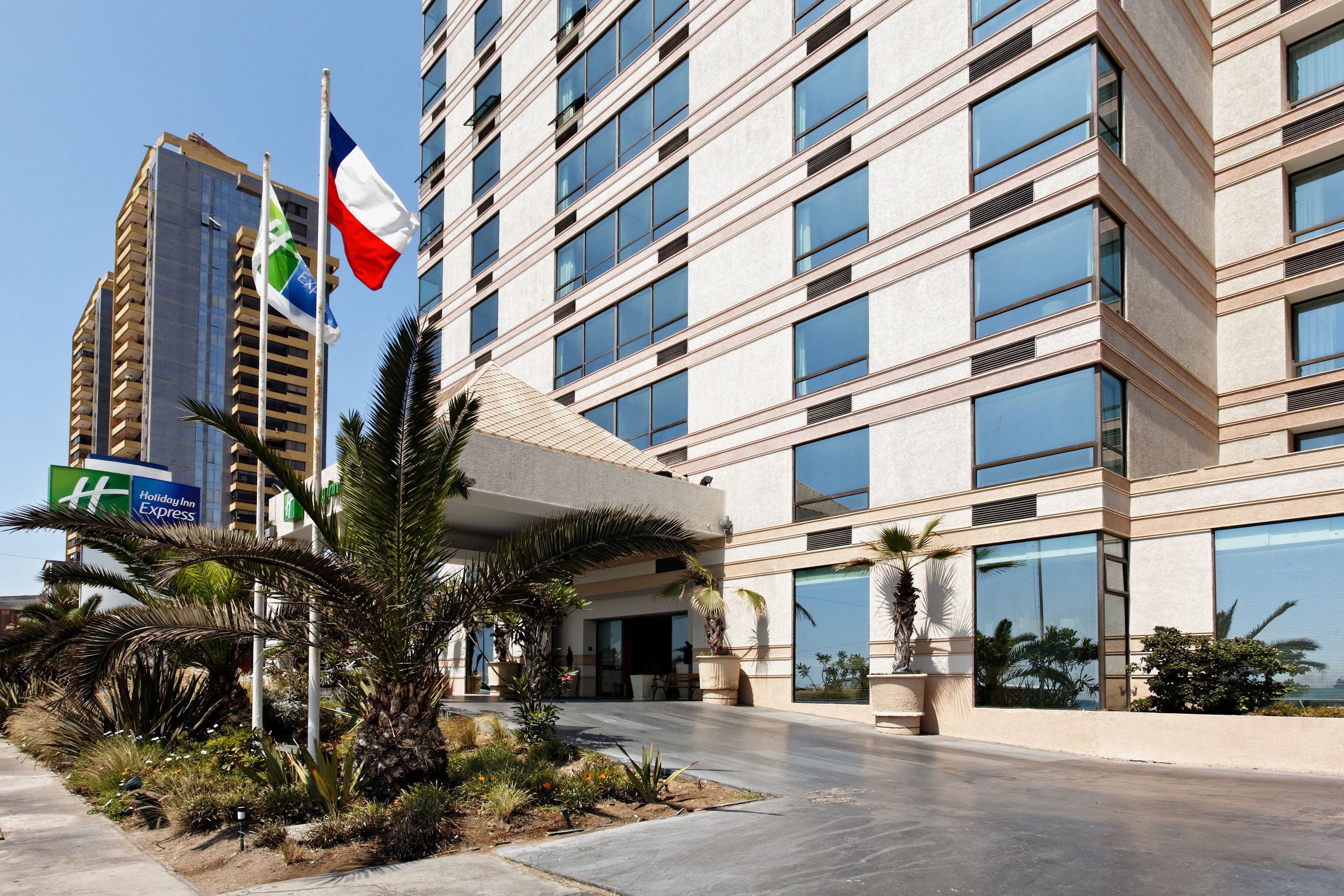 A multi-story hotel with a modern facade, large windows, and a landscaped entrance area with palm trees.