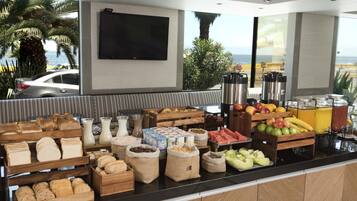 Enjoy a delicious breakfast spread with a variety of breads, pastries, fruits, and dairy products, served in a buffet area with a scenic ocean view.