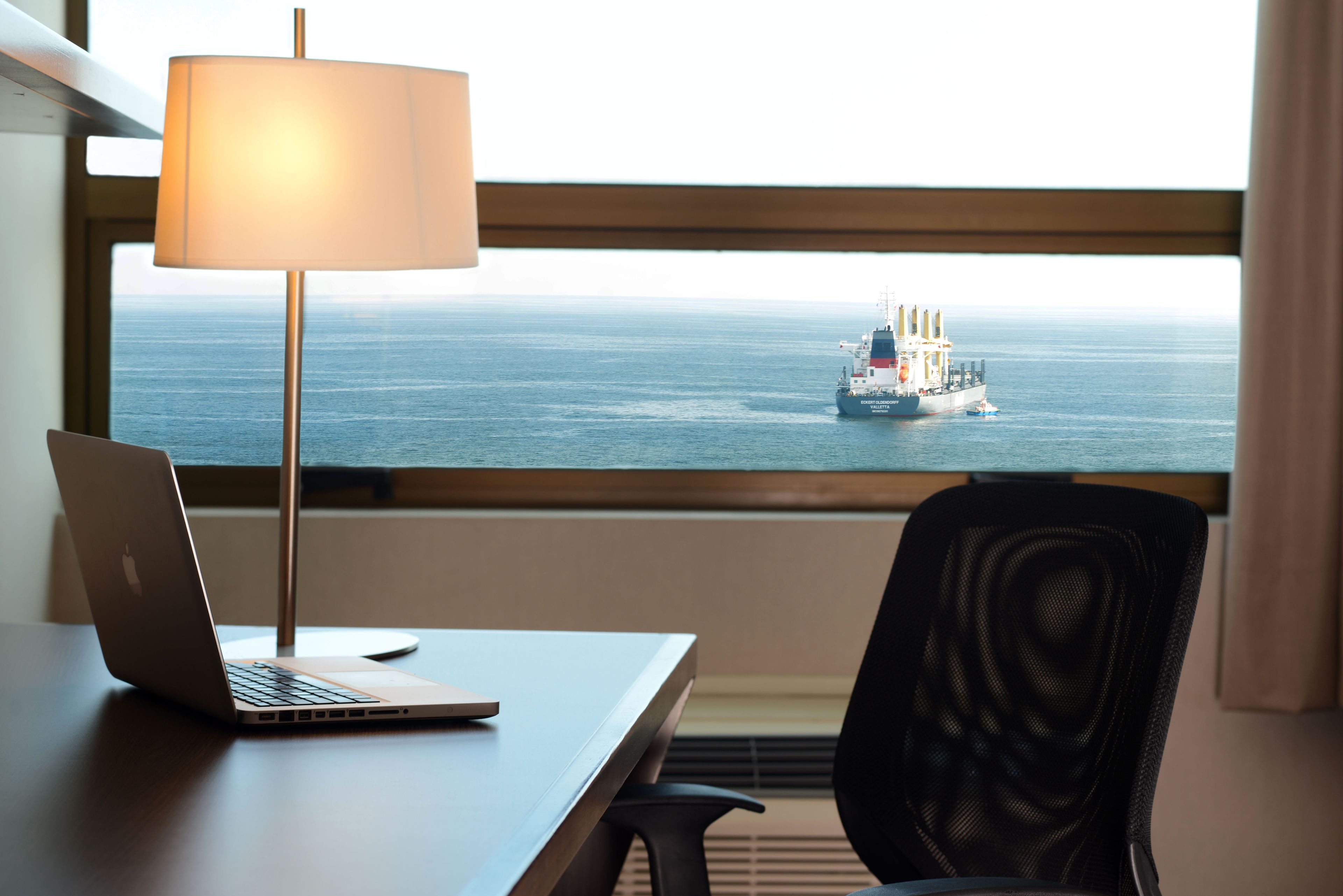 A laptop on a desk with a lamp, a chair, and a view of a ship on the water through a window.