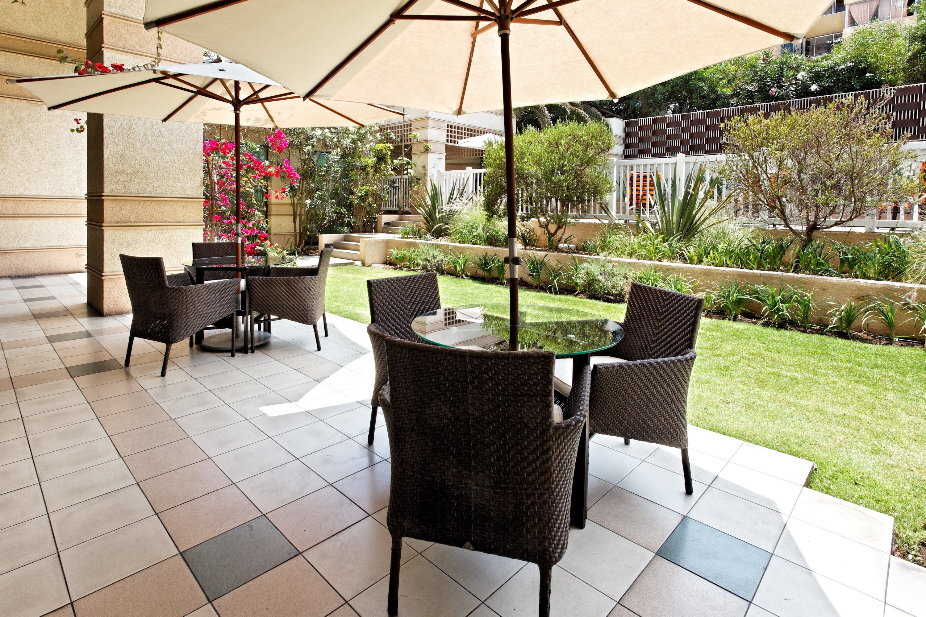 An outdoor patio area with wicker furniture and a glass table, surrounded by a well-maintained garden and a white fence.