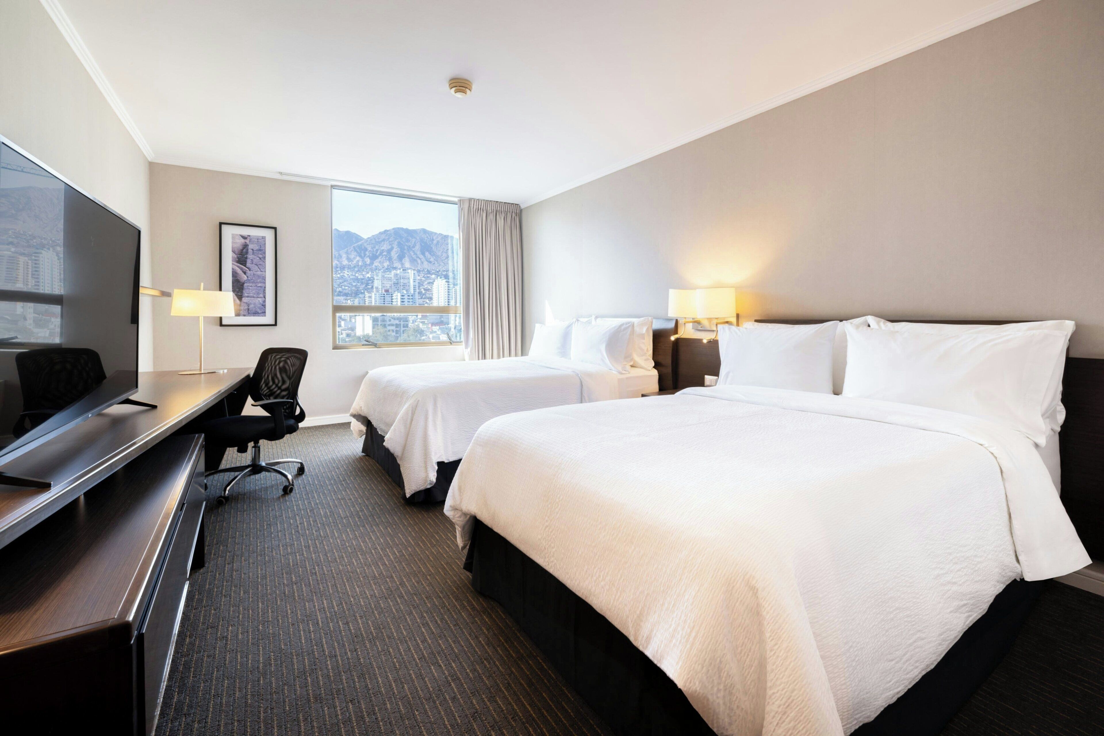 A hotel room with two beds, a desk, a chair, a TV, and a window with a city view.