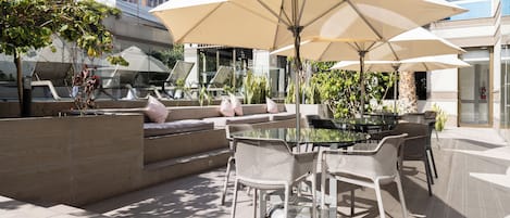 Un espace extérieur aménagé avec une table en verre, des chaises en osier blanc et des parasols beige.