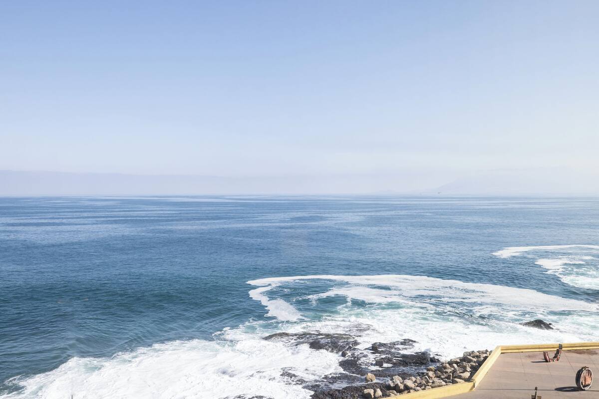 A coastal view with waves crashing against rocks, a clear sky, and a paved area with a railing.