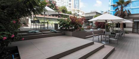 Un espacio al aire libre con escalones que conducen a una terraza, rodeado de vegetación y edificios modernos.