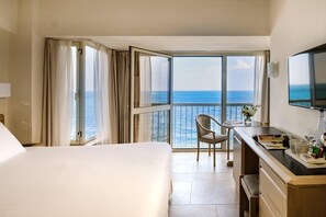 A hotel room with a bed, a desk with a chair, a TV, and a balcony with a view of the sea. - Diplomat Hotel (Sliema)