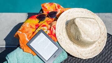 Un entorno frente al mar con un sombrero de paja trenzada, un par de gafas de sol, una toalla colorida y un dispositivo Kindle en una silla de mimbre junto a una piscina.