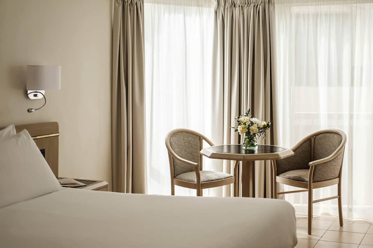 Una habitación de hotel con cama, mesita de noche, lámpara, silla, mesa y ventana con cortinas.