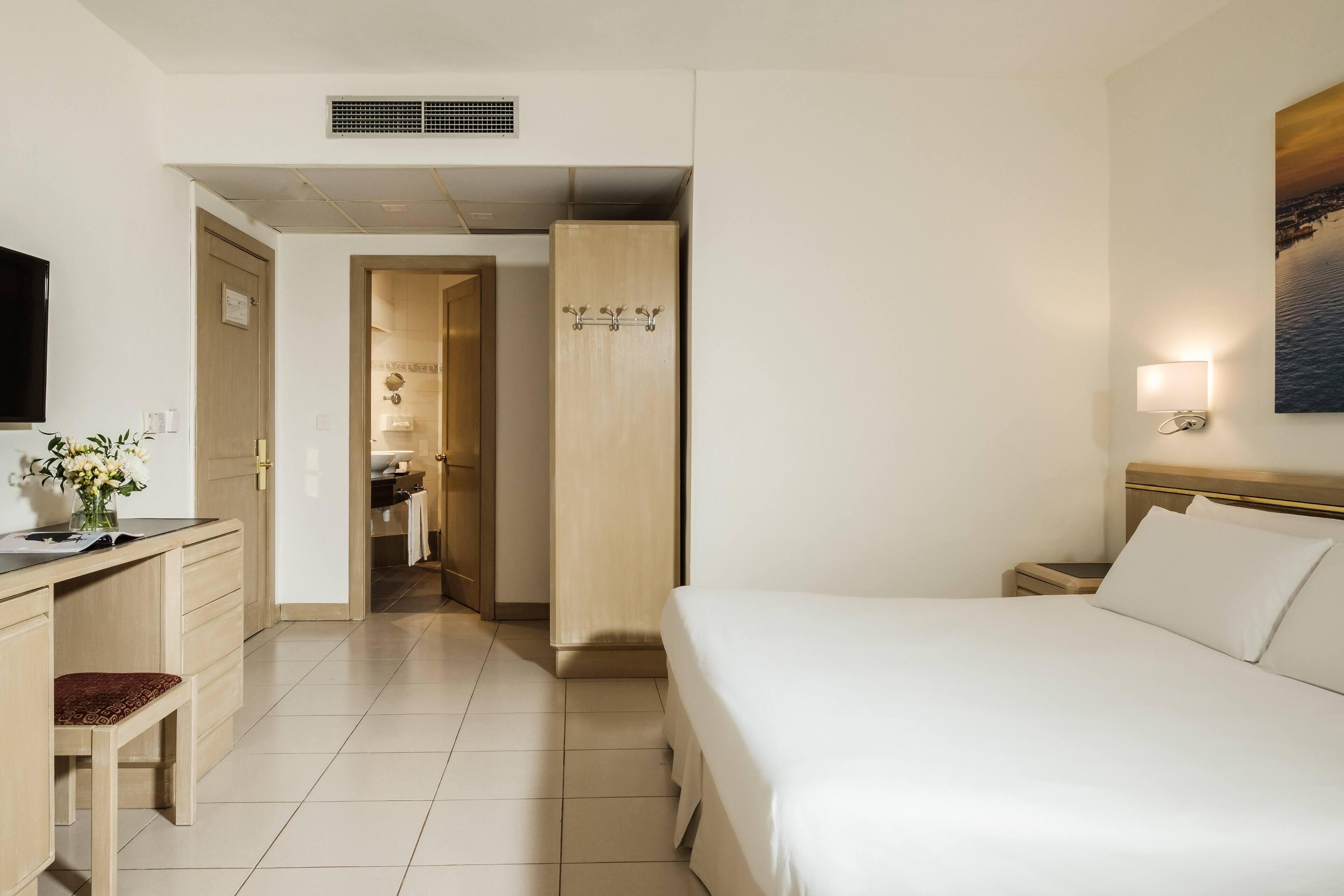 A hotel room with a bed, a desk with a chair, a wardrobe, a bathroom visible through an open door, and a painting on the wall.