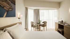 Habitación de hotel con cama, escritorio, silla, mesa y un ventanal grande con cortinas.