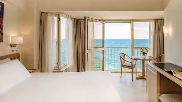 Habitación de hotel con cama, escritorio, silla y balcón con vista al mar.