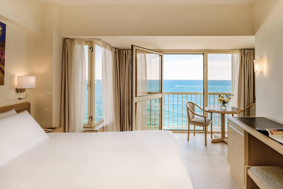 Habitación de hotel con cama, escritorio, silla y balcón con vista al mar.