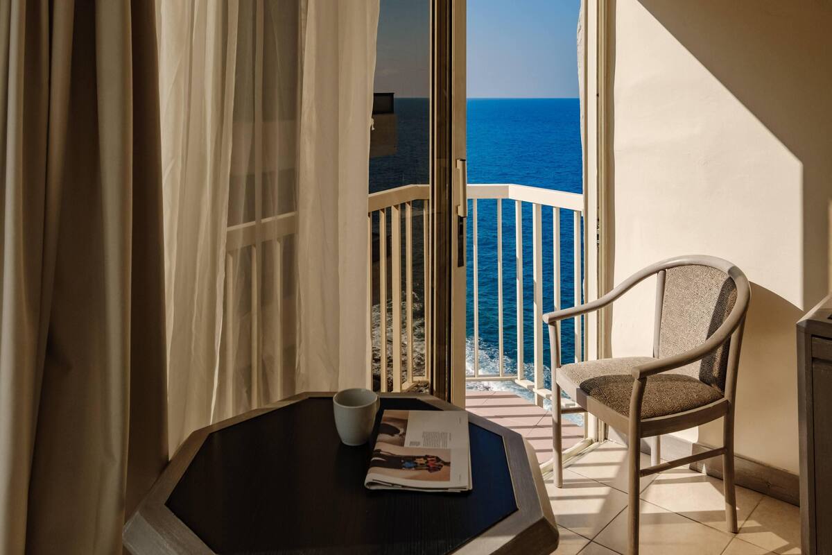 Un balcón con vista al mar, una silla, una mesa con una revista y una taza, y cortinas translúcidas.