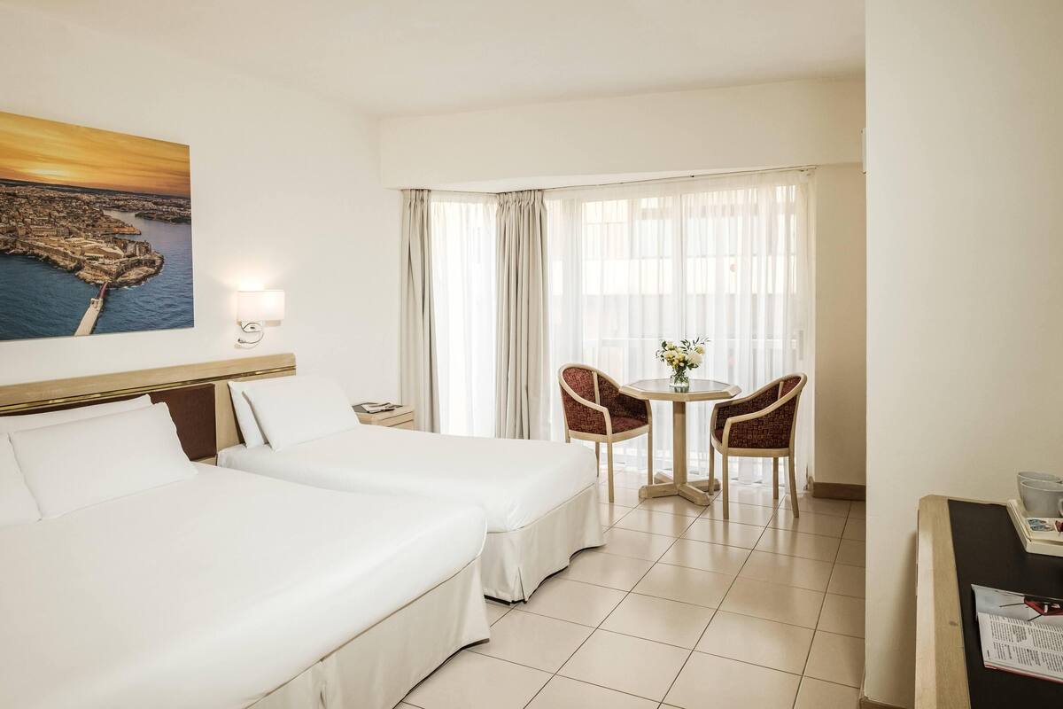 Habitación de hotel con dos camas, una mesa de comedor con sillas, un cuadro de un paisaje urbano costero y una ventana con cortinas ligeras.