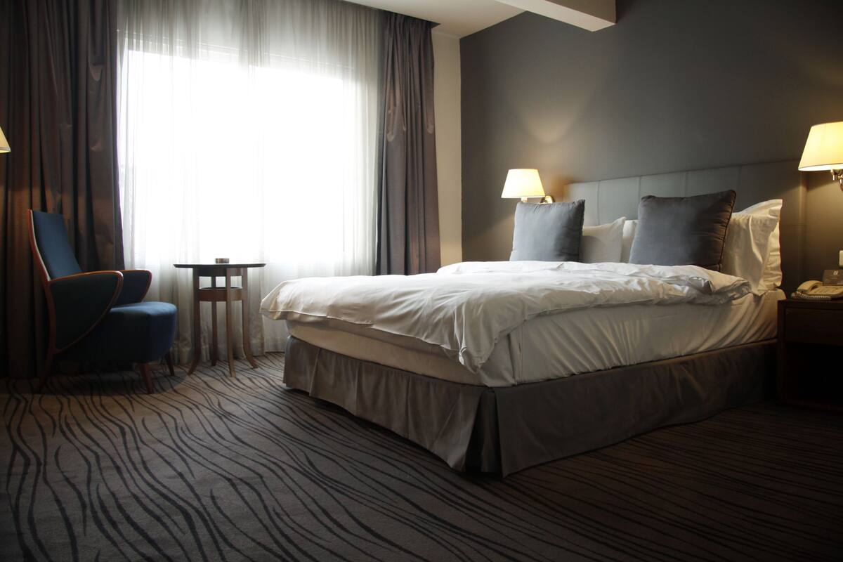 A hotel room with a large bed, two bedside lamps, a chair, a small table, and a view of the outdoors through sheer curtains.