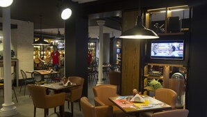 A restaurant with tables and chairs, a television screen, and a person in a red shirt working at the counter. - Plaza Hotel (Beirut)