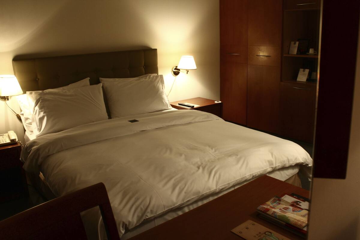 A neatly made bed with white linens, a bedside lamp, and a nightstand with a phone.