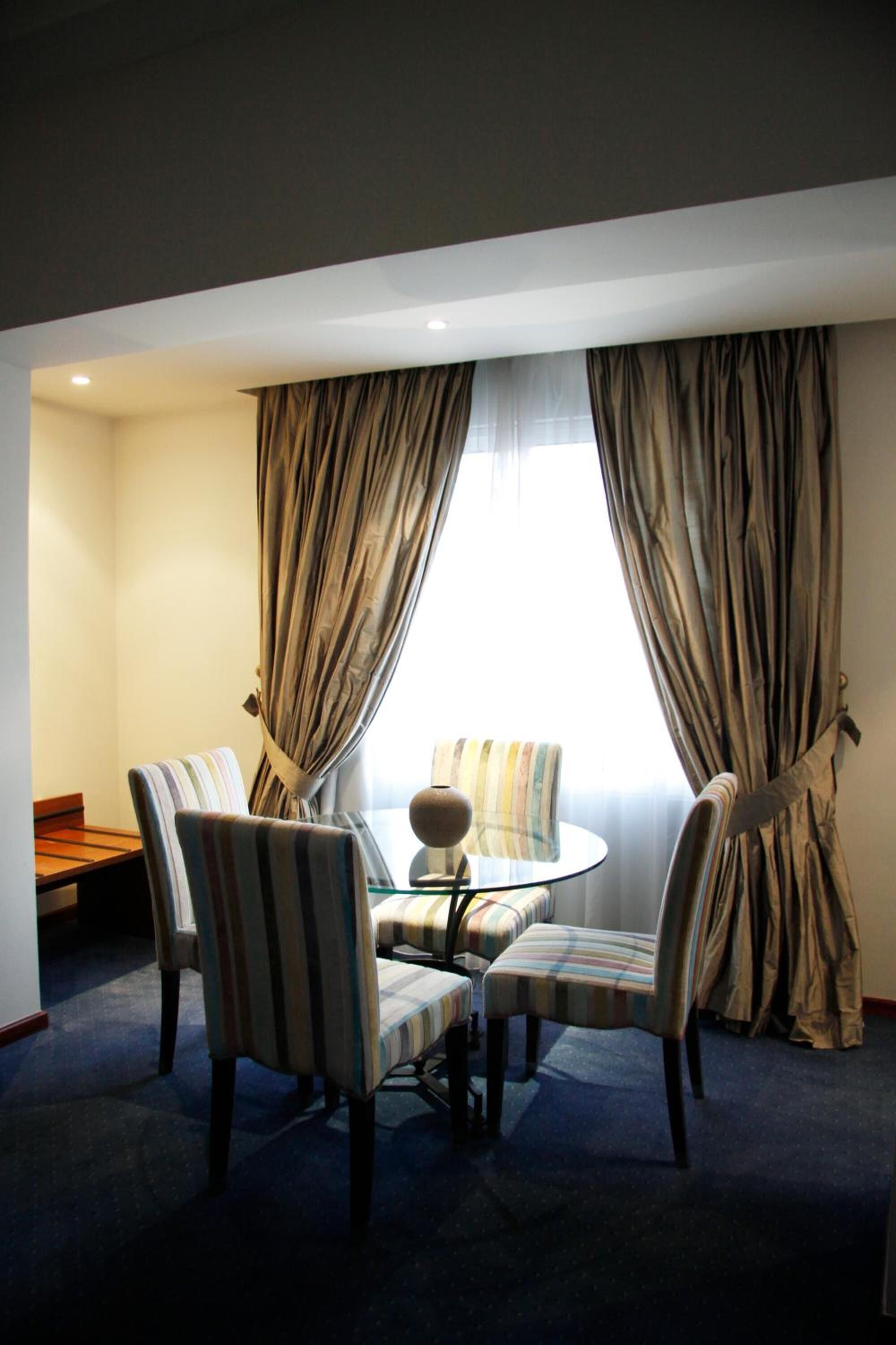 Habitación de hotel con zona de comedor que incluye sillas a rayas y una mesa redonda, un sofá y una ventana con cortinas.