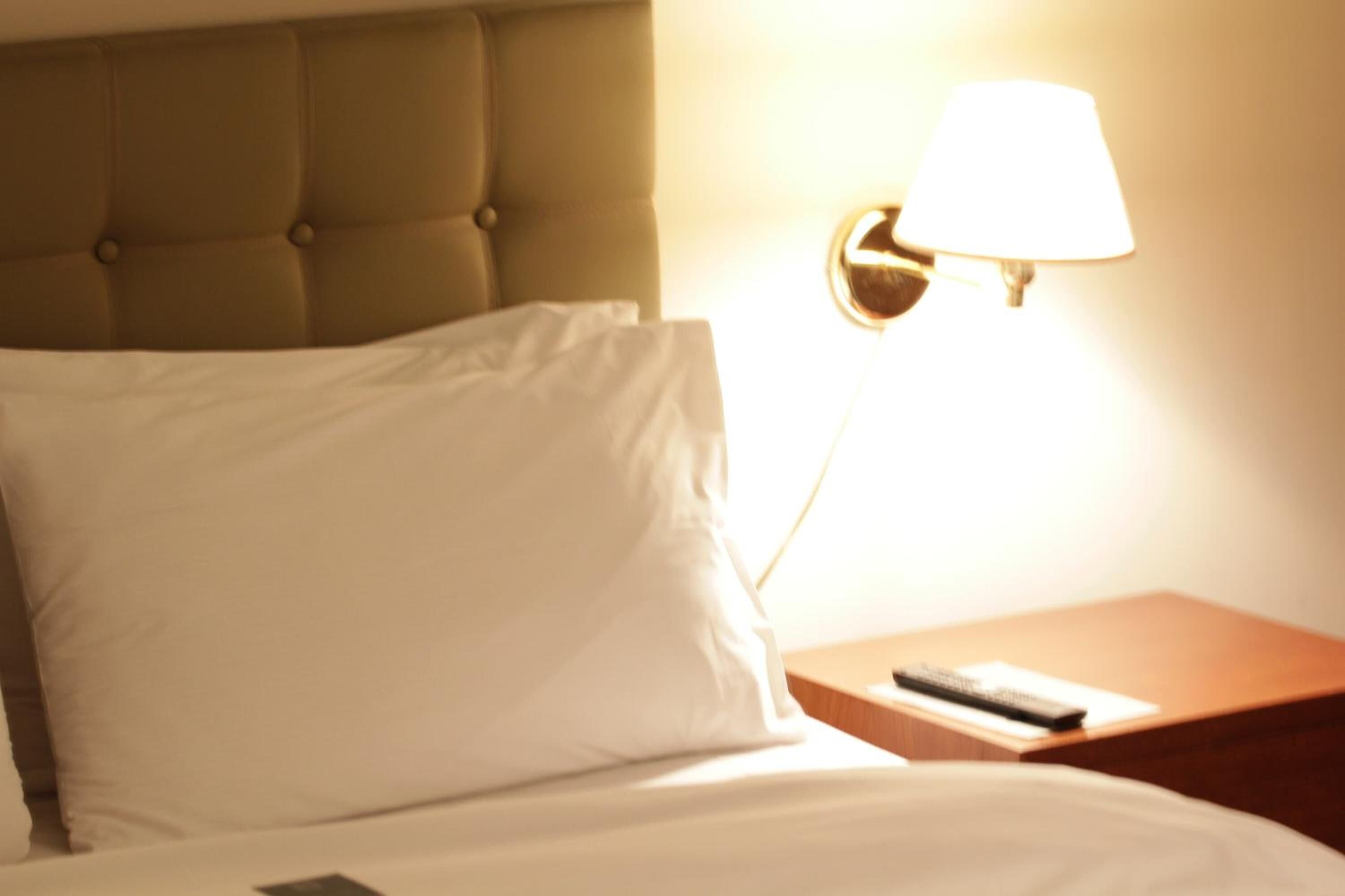 A neatly made bed with a white pillow and a remote control on a wooden nightstand beside it.