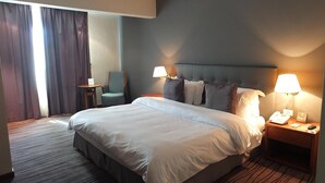 A hotel room with a large bed, two bedside lamps, a chair, a small table, and a nightstand with a phone and a sign. - Plaza Hotel (Beirut)