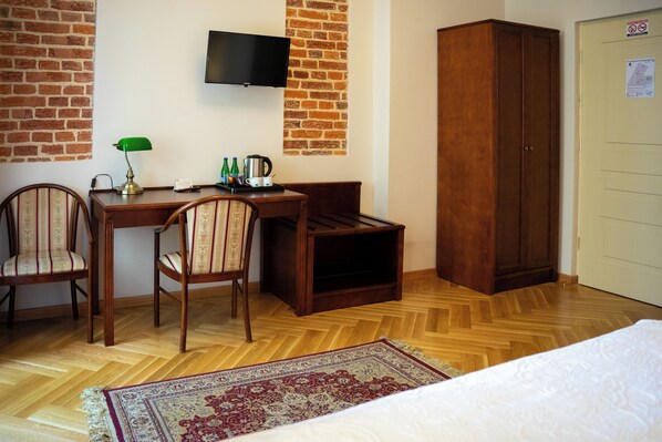 Habitación de hotel con un escritorio de madera, dos sillas, un televisor montado en una pared de ladrillo, un armario y una alfombra estampada en el suelo.