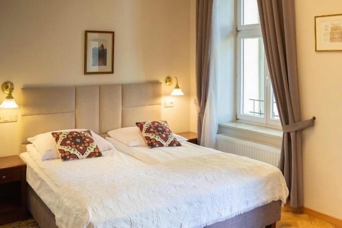 Una cama doble con edredón blanco, dos mesitas de noche con lámparas, una ventana con cortinas y un cuadro en la pared.