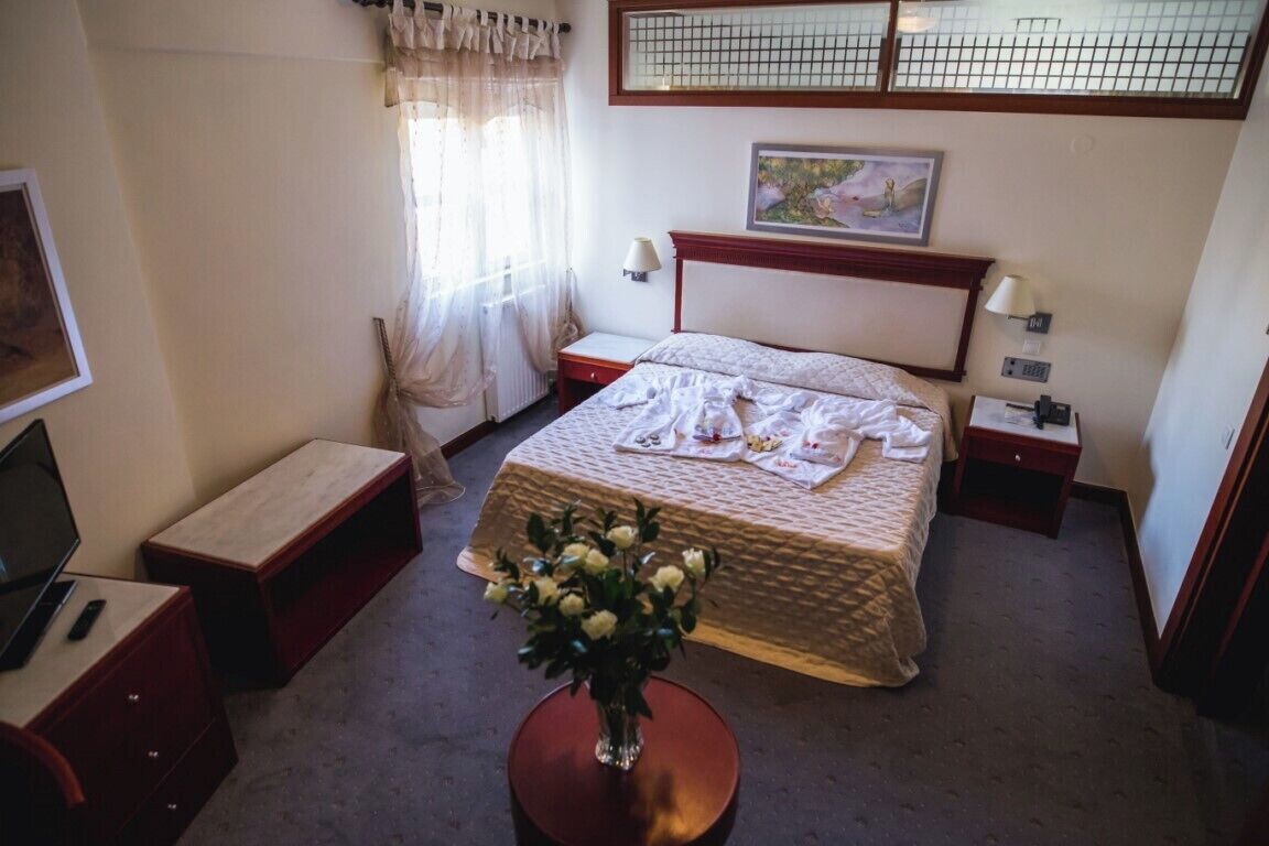 A hotel room with a bed, bedside tables, a desk with a chair, a vase with flowers, and a window with curtains.
