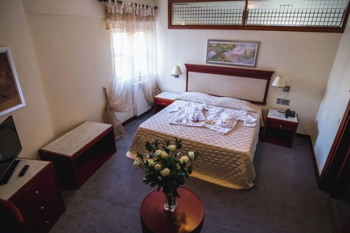 A hotel room with a bed, bedside tables, a desk with a chair, a vase with flowers, and a window with curtains.