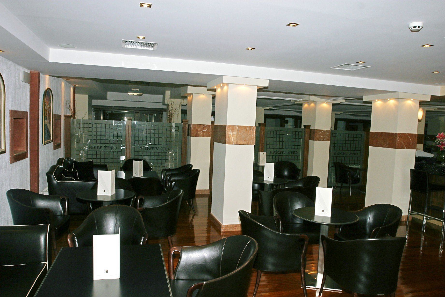 Un hall d’hôtel doté de plusieurs espaces aménagés avec des sièges, notamment des fauteuils et des tables en cuir noir. On y trouve également des piliers ornés d’éléments décoratifs et une cloison en verre portant une inscription.