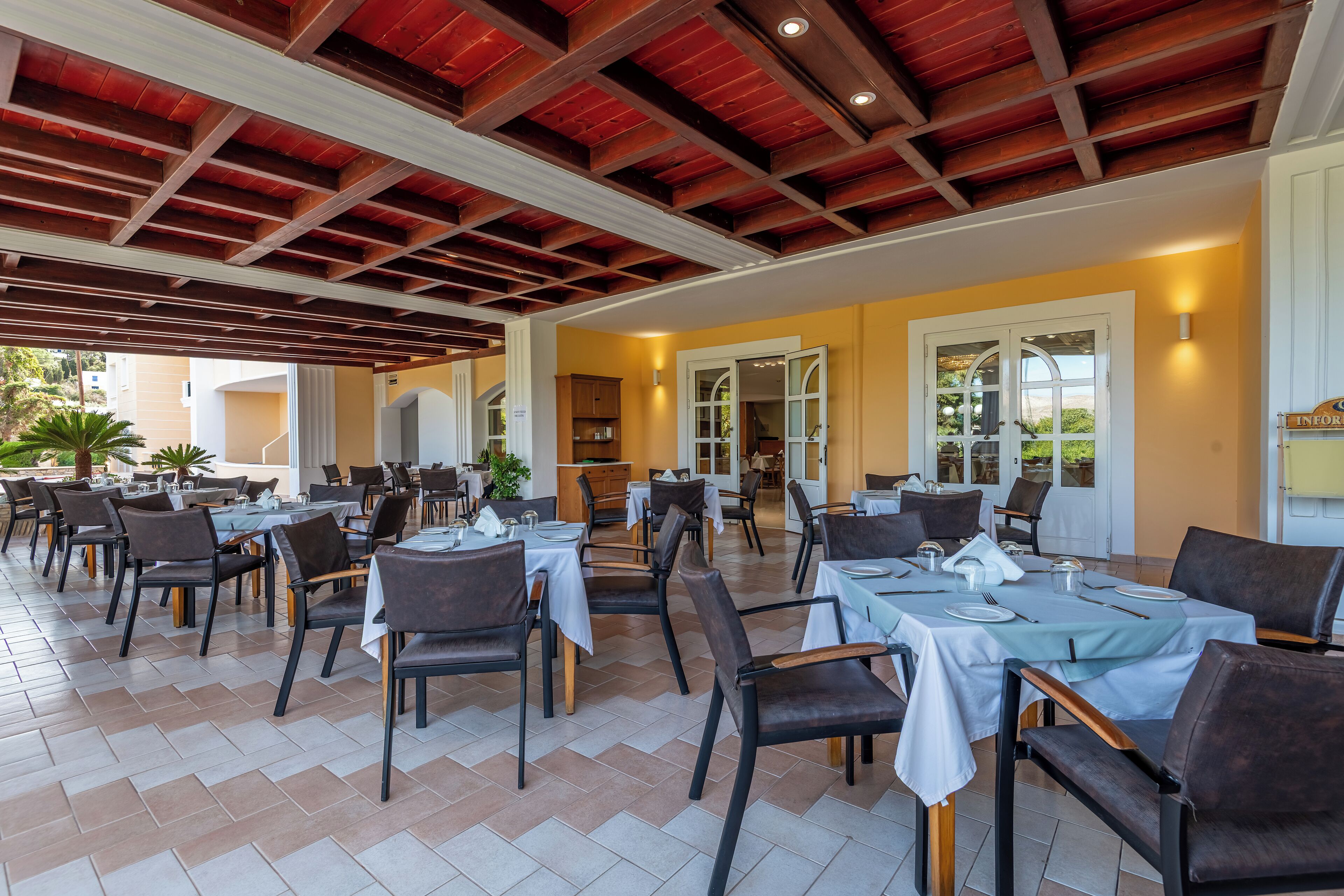 An outdoor dining area with multiple tables and chairs, wooden ceiling beams, and a tiled floor.