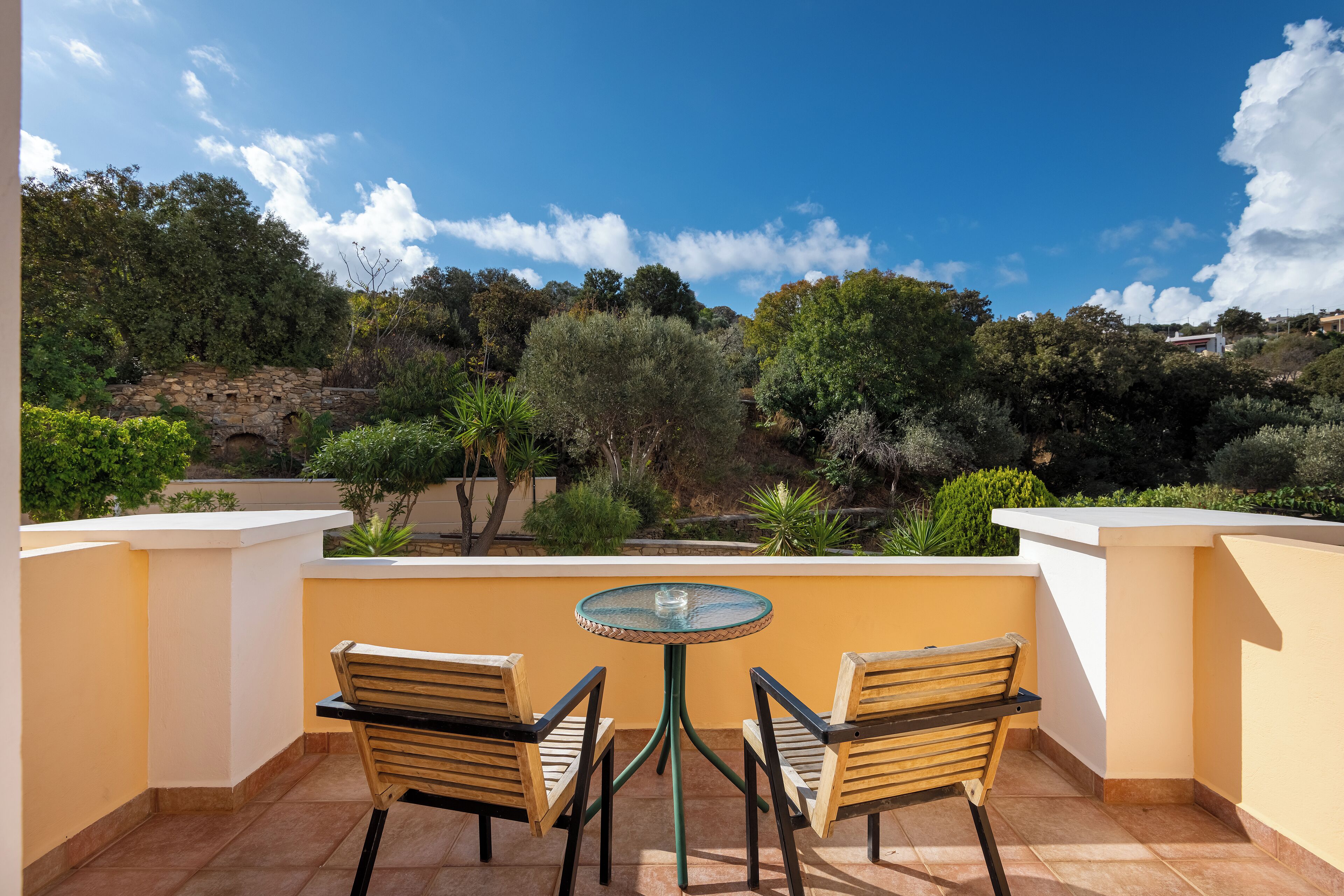 A balcony with two wooden chairs and a glass table, overlooking a landscaped garden with various trees and shrubs.