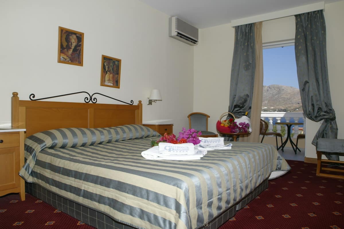 A hotel room with a bed, a nightstand, a lamp, a chair, a table with a vase of flowers, and a window with curtains.