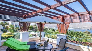 Un balcón con una silla reclinable verde, una mesa con una botella y vasos, y vistas al mar y las colinas.