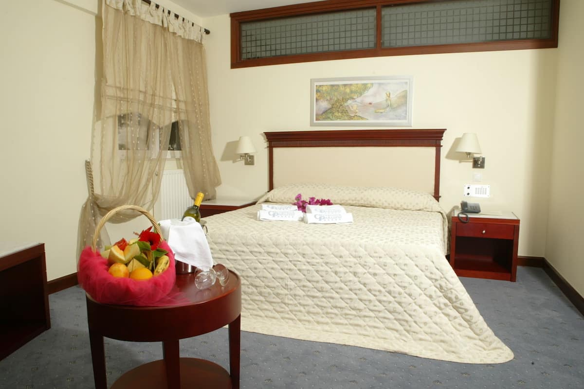 A hotel room with a large bed, a bedside table with a lamp, a nightstand, a window with curtains, and a wall-mounted artwork.