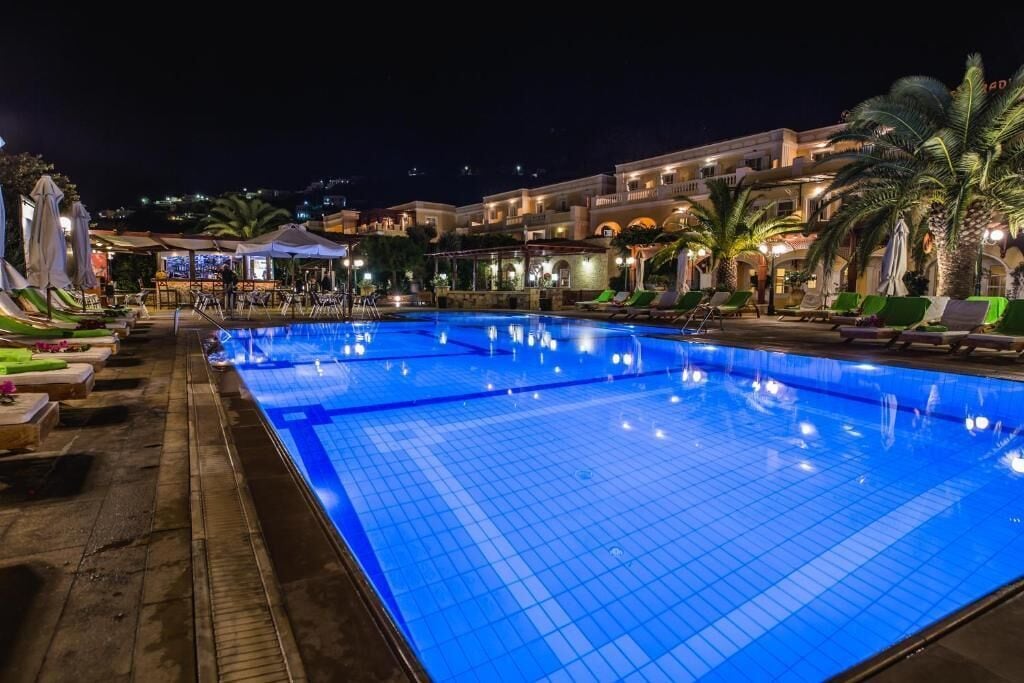 A swimming pool with lounge chairs and umbrellas, surrounded by buildings with balconies and palm trees.