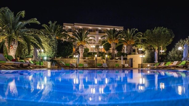 Una piscina con sillas de descanso y palmeras, rodeada por un edificio con varios balcones e iluminación.
