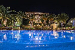 A swimming pool with lounge chairs and palm trees, surrounded by a building with multiple balconies and lights.