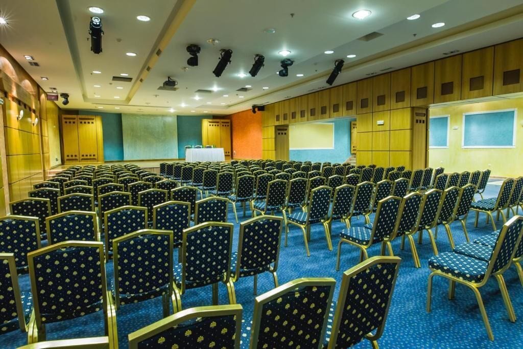 A conference room with rows of chairs, a stage with a podium, and a large screen.