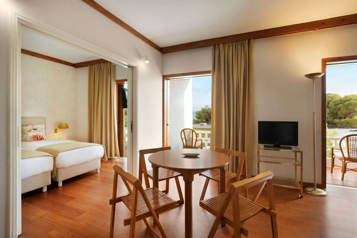 A hotel room with a bed, a dining table with chairs, a TV on a stand, and large windows with curtains.