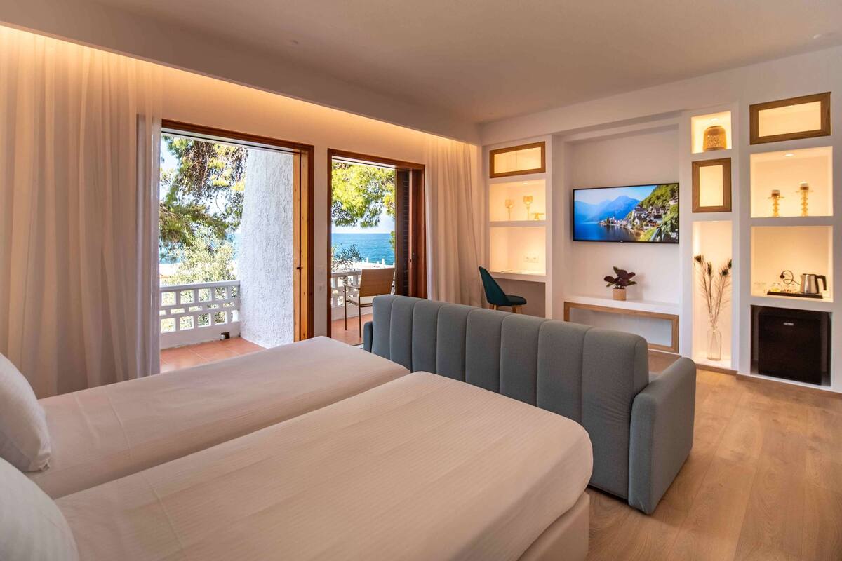 A hotel room with a large bed, a sofa, a TV, and a balcony with a view of the sea.