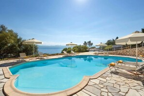 Ein Pool mit Liegestühlen und Sonnenschirmen, umgeben von Steinplatten und Blick aufs Meer.