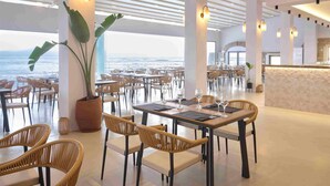 A spacious dining area with high ceilings, large windows, and a view of the ocean. Tables are set with wine glasses and napkins. - Wyndham Loutraki Poseidon Resort (Loutraki-Agioi Theodoroi)
