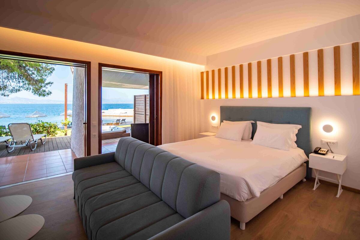 A modern bedroom with a large bed, a sofa, and a view of the sea through an open door.