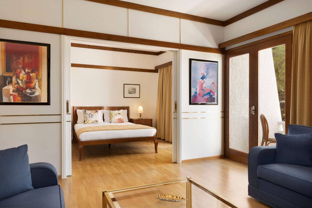 A modern hotel room with a wooden floor, a bed with a beige headboard, two blue sofas, a glass coffee table, and artwork on the walls.