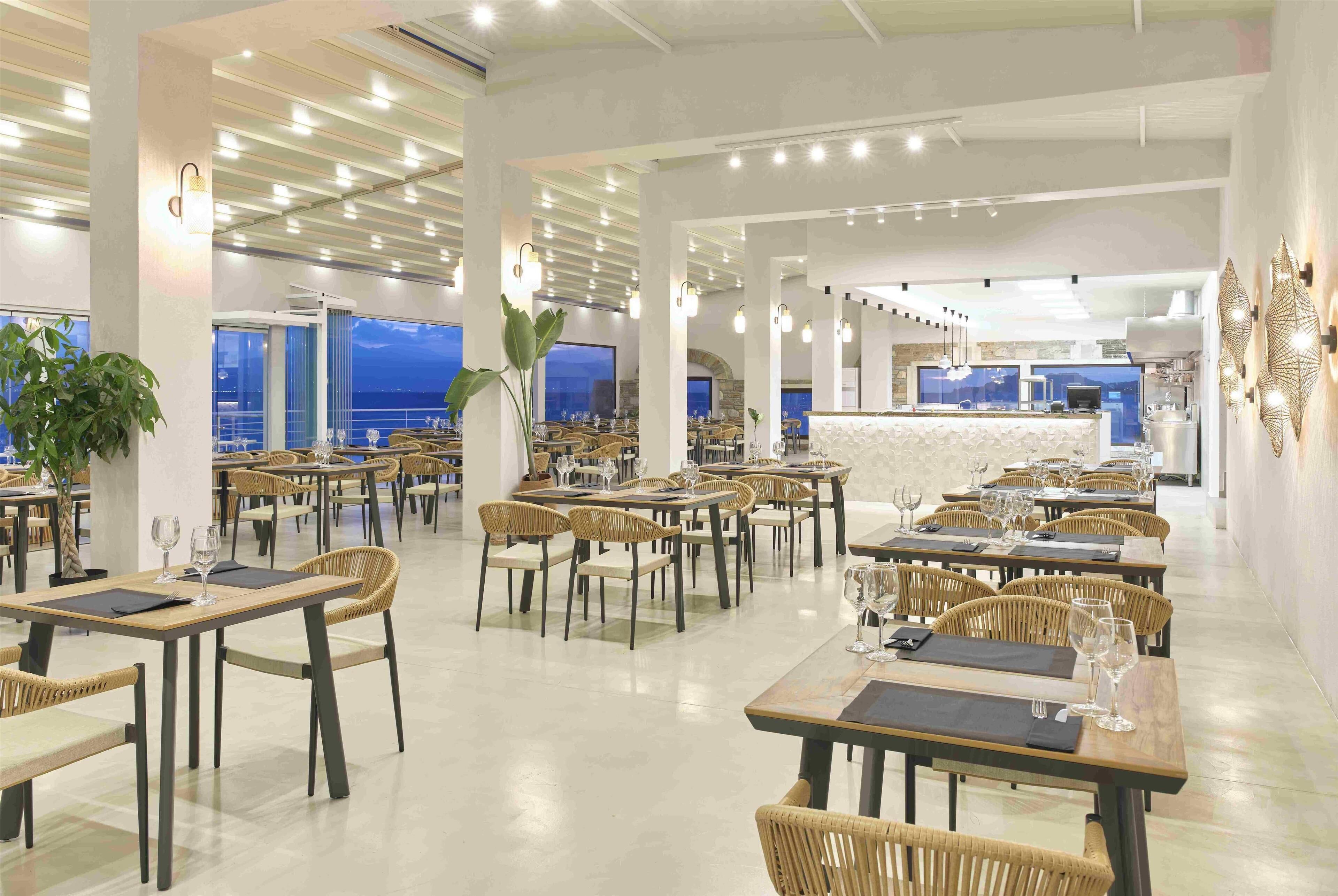 A spacious dining area with multiple tables and chairs, a central bar area, and large windows offering views of the sea.