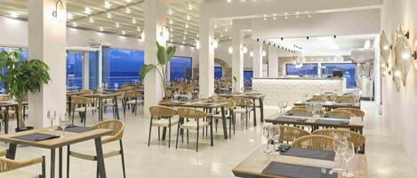 A spacious dining area with multiple tables and chairs, a central bar area, and large windows offering views of the sea.