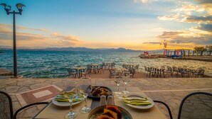 A dining area with tables and chairs overlooking a body of water, a diving boat, and a distant mountain range. - Wyndham Loutraki Poseidon Resort (Loutraki-Agioi Theodoroi)