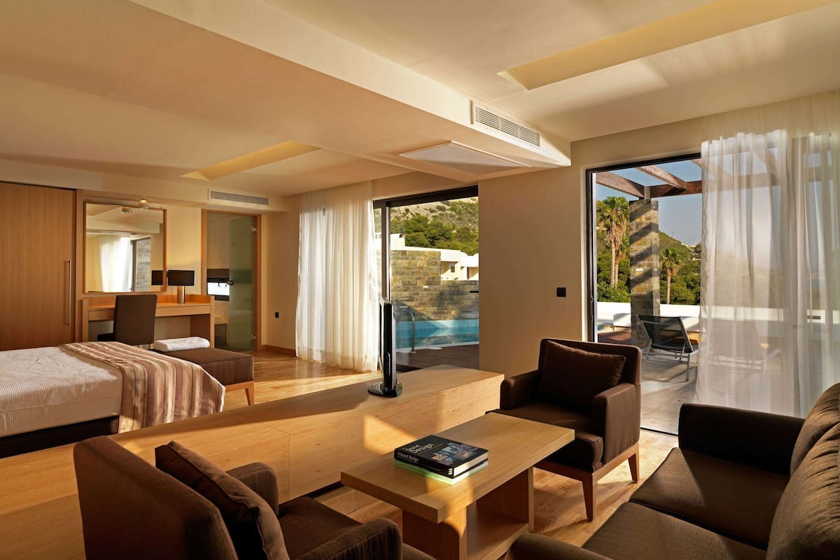 A modern hotel room with a bed, a desk, a sofa, and a view of a pool and mountains.